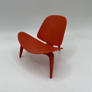 Może przedstawiać: Miniaturowy czerwony fotel z zakrzywionym oparciem i czterema nogami. Fotel wykonany jest z gładkiego, plastikowego materiału.