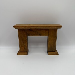 May include: A small, wooden fireplace mantel in a medium brown color. The mantel has a rectangular top and two vertical supports with a rectangular base. The design is simple and classic, suitable for a dollhouse or miniature setting.