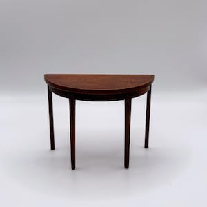 Puede incluir: Pequeña mesa de madera en forma de semicírculo con acabado marrón oscuro. Tiene cuatro patas delgadas y rectas y una superficie curva y lisa. La mesa mide aproximadamente 61 cm de ancho y 30 cm de profundidad.