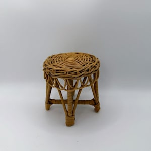 May include: A miniature stool crafted from light brown woven wicker. The seat features a circular, tightly coiled pattern, while the base consists of four slender legs connected by horizontal support struts in a simple, open-frame design.