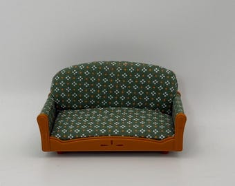 May include: A miniature sofa featuring a solid orange plastic frame with a curved back and armrests. The seat and backrest are upholstered in a dark green fabric patterned with a repeating design of small white four-petaled flowers and orange dots.