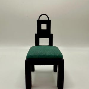 May include: A small, black wooden chair with a square seat cushion in a dark green color. The chair has a unique design with a square cut-out in the back and a small handle at the top. The chair is a minimalist design.
