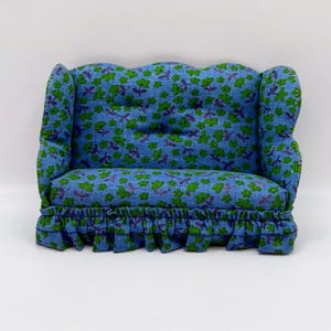 May include: A miniature blue sofa with green floral patterns. The sofa has a ruffled skirt and curved armrests. The fabric is a light blue with a repeating pattern of green flowers. The sofa is designed for a dollhouse or miniature setting.