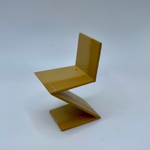 Può includere: Una sedia a zig-zag in miniatura di colore giallo realizzata in legno. La sedia ha un design semplice con una leggera curva nella seduta e nello schienale.