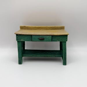 May include: A small, rustic-style wooden table with a green base and a light brown tabletop. The table features a single drawer with a dark handle. The wood grain is visible, giving it a textured appearance. The table is designed with a simple, functional aesthetic.