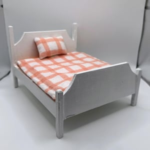 May include: A miniature white bed with a headboard and footboard. The bed is made of wood and has a peach and white plaid blanket and pillow. The bed is a dollhouse accessory.