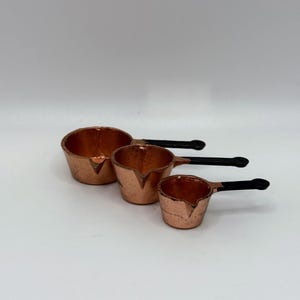 May include: Three copper-colored saucepans with black handles are arranged on a white surface. The saucepans vary in size, with the largest on the left and the smallest on the right. The handles are attached with rivets.