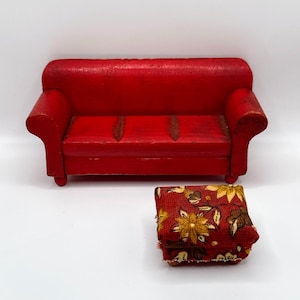 May include: A miniature red wooden sofa with a matching patterned ottoman. The sofa has a classic design with rolled arms and a three-cushion seat. The ottoman is a small, square shape with a floral pattern in shades of yellow, brown, and cream.