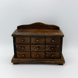 May include: A small, dark brown wooden chest with a rectangular shape and a curved top edge. The chest features nine individual drawers arranged in three rows of three, each equipped with a tiny, centered round knob for opening and closing.