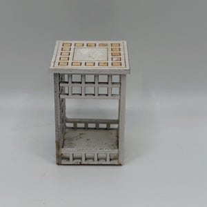 Op de afbeelding: Een kleine, witte decoratieve tafel met een vierkant blad en een roosterachtig ontwerp. De tafel heeft een rechthoekige vorm met een plank en roosterachtige zijkanten. De bovenkant heeft een centraal vierkant omgeven door kleinere vierkanten.