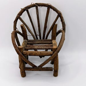 May include: A miniature chair constructed from dark brown, textured twigs and branches. The backrest features a fan-like arrangement of vertical sticks within a circular frame, while the seat and legs are made of straight, horizontal wooden pieces.