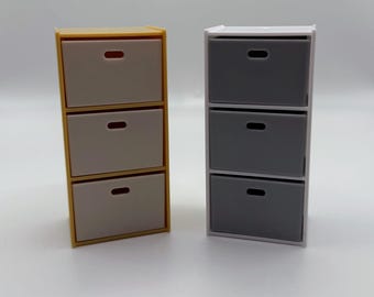 Puede incluir: Dos mini-cómodas de tres cajones. Una cómoda tiene un marco amarillo con cajones gris claro. La otra cómoda tiene un marco blanco con cajones grises. Cada cajón tiene un pequeño tirador horizontal.