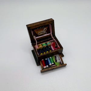 May include: A miniature wooden sewing box with an open top and drawer. The box contains spools of thread in various colors, scissors, buttons, and a measuring tape. The box is brown with a pink interior.