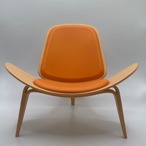 Może przedstawiać: Krzesło z jasnego drewna z zakrzywionym oparciem i jasno pomarańczową poduszką. Krzesło ma unikalny, nowoczesny design.