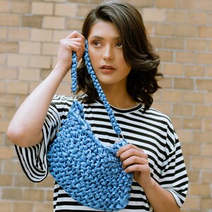 May include: A blue handbag featuring a textured, woven surface and a braided strap attached with silver-toned metal rings. The bag is constructed from a continuous, knotted material, creating a dense, repetitive pattern throughout the body.