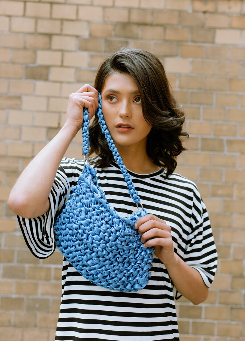 May include: A blue handbag featuring a textured, woven surface and a braided strap attached with silver-toned metal rings. The bag is constructed from a continuous, knotted material, creating a dense, repetitive pattern throughout the body.