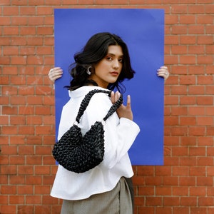 May include: A black woven shoulder bag with a braided strap and circular metal rings, featuring a textured, knotted surface design. The bag is compact with a curved silhouette and is constructed from a dark, glossy material.