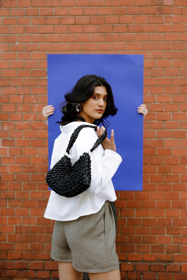 May include: A black woven shoulder bag with a braided strap and circular metal rings, featuring a textured, knotted surface design. The bag is compact with a curved silhouette and is constructed from a dark, glossy material.