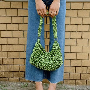 May include: A bright lime green handbag crafted from woven satin ribbon, featuring a braided handle attached with gold-toned metal rings. The bag has a textured, knotted surface and a slouchy, crescent-shaped silhouette.