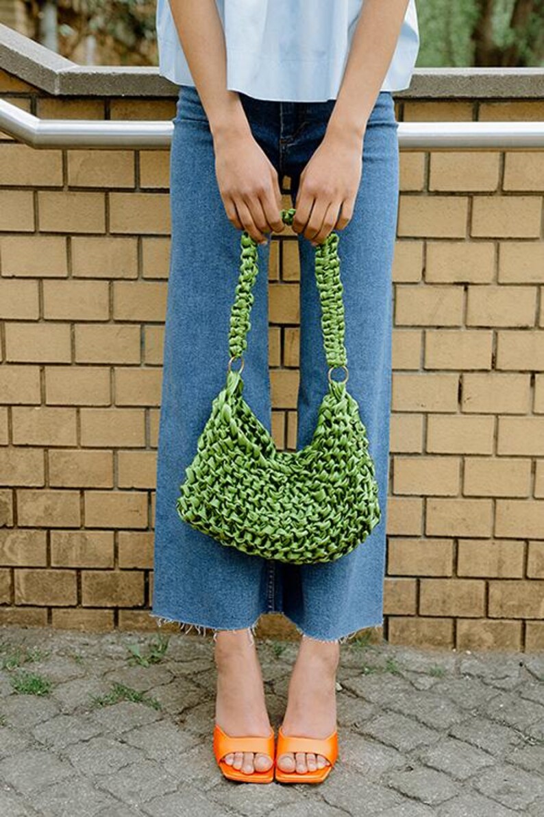 May include: A bright lime green handbag crafted from woven satin ribbon, featuring a braided handle attached with gold-toned metal rings. The bag has a textured, knotted surface and a slouchy, crescent-shaped silhouette.