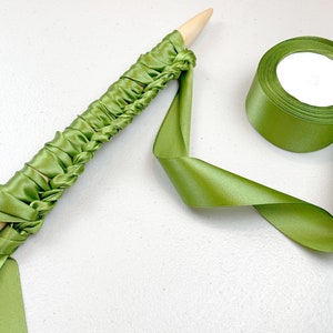 May include: A light wooden knitting needle wrapped in a continuous length of olive green satin ribbon, featuring a series of gathered loops along the shaft, accompanied by a matching spool of olive green satin ribbon.