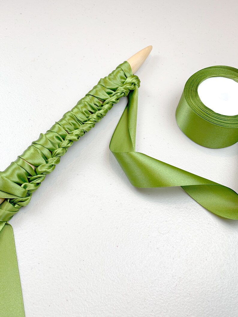 May include: A light wooden knitting needle wrapped in a continuous length of olive green satin ribbon, featuring a series of gathered loops along the shaft, accompanied by a matching spool of olive green satin ribbon.