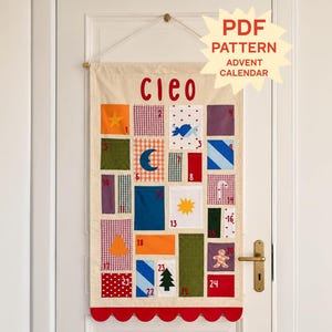 May include: A hanging fabric advent calendar with the name Cleo in red letters at the top. It features twenty-four numbered pockets in various colors and patterns, including gingham, polka dots, and solid blocks, with festive icons like a star, moon, sun, and tree.