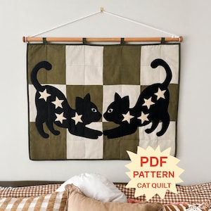 Puede incluir: Un edredón de pared con un patrón de tablero de ajedrez de cuadrados verde oliva y crema. Dos siluetas de gatos negros con aplicaciones de estrellas color crema están centradas en el edredón. Una varilla de madera sostiene el edredón con una etiqueta PDF.