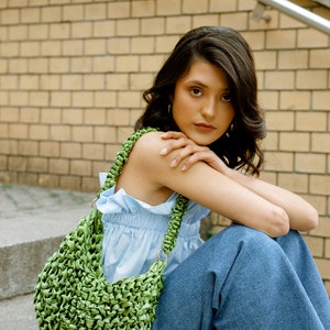 May include: A bright green woven handbag featuring a textured, knotted ribbon construction. The bag has a single shoulder strap attached with silver-toned metal rings, creating a slouchy, crescent-shaped silhouette with a dense, intricate pattern.