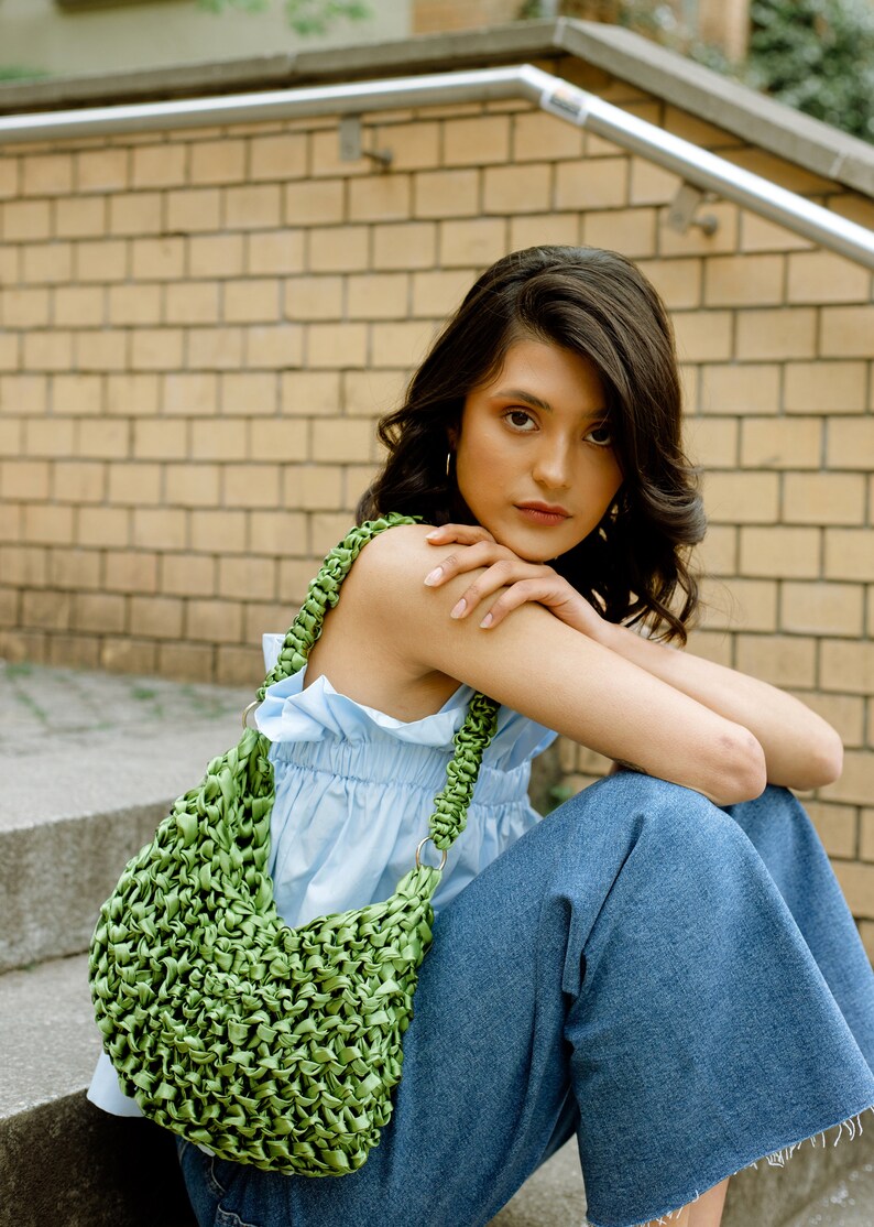 May include: A bright green woven handbag featuring a textured, knotted ribbon construction. The bag has a single shoulder strap attached with silver-toned metal rings, creating a slouchy, crescent-shaped silhouette with a dense, intricate pattern.