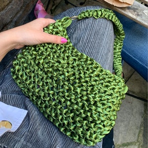 May include: A vibrant olive green handbag crafted from a thick, woven satin ribbon material. The bag features a rectangular body with a matching braided shoulder strap attached by a gold-toned metal ring on one side, showcasing a textured finish.