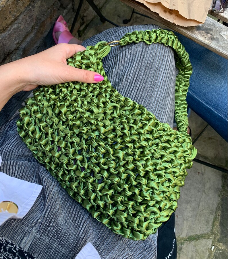 May include: A vibrant olive green handbag crafted from a thick, woven satin ribbon material. The bag features a rectangular body with a matching braided shoulder strap attached by a gold-toned metal ring on one side, showcasing a textured finish.