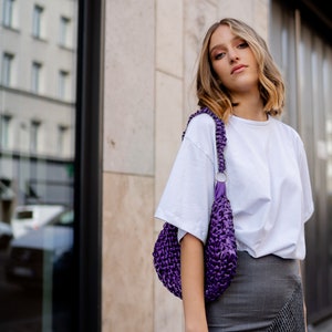 May include: A deep purple, hand-crocheted shoulder bag with a thick, braided strap and a silver-toned metal ring connector, worn over a plain white short-sleeved t-shirt and a grey, plaid-patterned skirt with a contrasting textured panel.