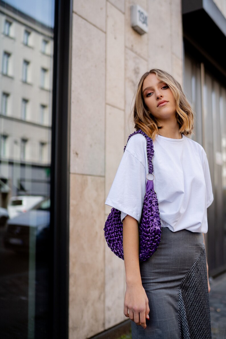 May include: A deep purple, hand-crocheted shoulder bag with a thick, braided strap and a silver-toned metal ring connector, worn over a plain white short-sleeved t-shirt and a grey, plaid-patterned skirt with a contrasting textured panel.