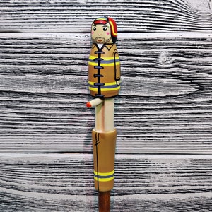 May include: A wooden figurine of a firefighter wearing a yellow and black uniform with a red helmet. The figurine is standing on a wooden base.