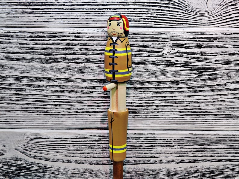 May include: A wooden figurine of a firefighter wearing a yellow and black uniform with a red helmet. The figurine is standing on a wooden base.