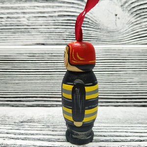 Pode incluir: Uma figura de madeira de um bombeiro vestindo um capacete vermelho e um uniforme listrado preto e amarelo. A figura &eacute; pintada com detalhes e tem uma fita vermelha presa ao topo do capacete.