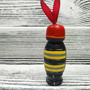 Pode incluir: Uma boneca de madeira com um corpo listrado preto e amarelo, um chap&eacute;u vermelho com uma faixa dourada e uma fita vermelha no topo. A boneca est&aacute; em p&eacute; sobre uma superf&iacute;cie de madeira branca.