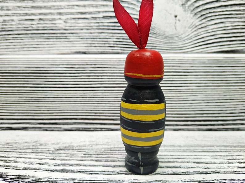 Pode incluir: Uma boneca de madeira com um corpo listrado preto e amarelo, um chap&eacute;u vermelho com uma faixa dourada e uma fita vermelha no topo. A boneca est&aacute; em p&eacute; sobre uma superf&iacute;cie de madeira branca.