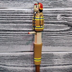 May include: A wooden figurine of a firefighter wearing a red helmet and a yellow and black uniform. The figurine is painted with details, including a mustache and a friendly face.