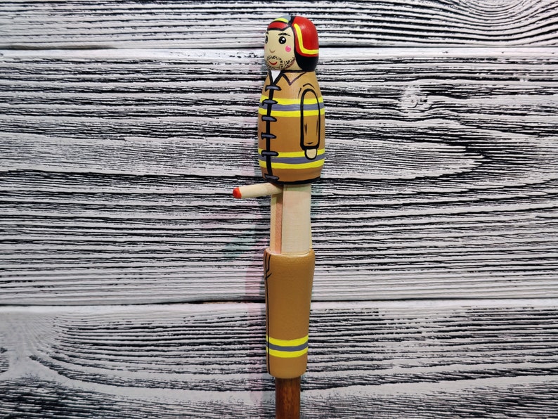 May include: A wooden figurine of a firefighter wearing a red helmet and a yellow and black uniform. The figurine is painted with details, including a mustache and a friendly face.