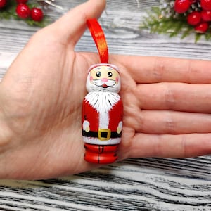 可能包含:手绘木制圣诞老人圣诞树挂饰。挂饰造型小巧,身穿红色外套,配以白色毛皮饰边和黑色腰带。挂饰上系着一条红丝带,面容欢快,脸颊红润。这款迷人的挂饰非常适合为您的节日装饰增添一抹手工奇思妙想。