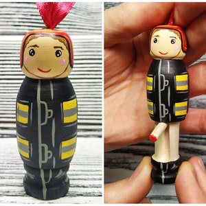 Pode incluir: Um ornamento de boneca de madeira pintado para parecer um bombeiro. A boneca est&aacute; vestindo um traje de bombeiro preto e amarelo com um capacete vermelho. A boneca tem um rosto pintado com cabelo castanho e um sorriso.