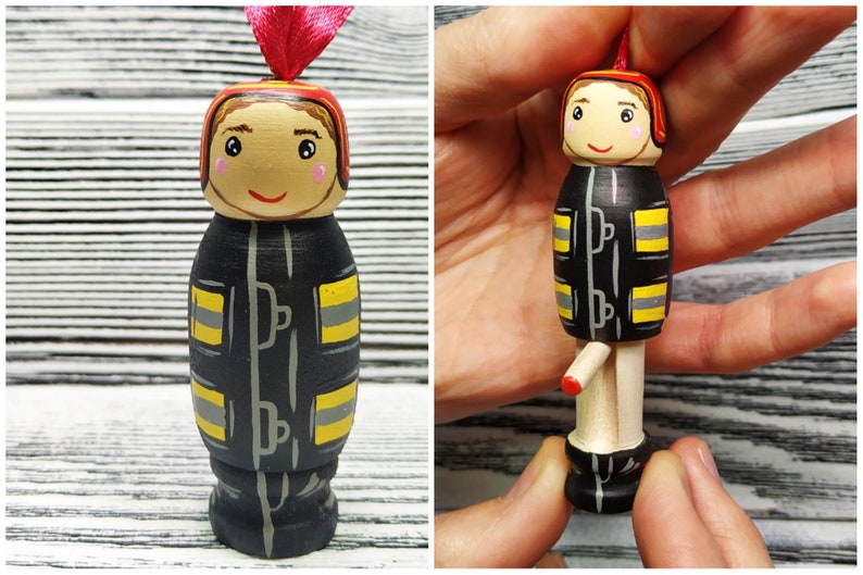 Pode incluir: Um ornamento de boneca de madeira pintado para parecer um bombeiro. A boneca est&aacute; vestindo um traje de bombeiro preto e amarelo com um capacete vermelho. A boneca tem um rosto pintado com cabelo castanho e um sorriso.