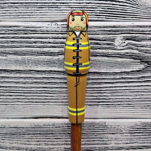 May include: A wooden figurine of a firefighter wearing a brown uniform with yellow reflective stripes and a red helmet. The figurine has a painted face with a mustache and a friendly expression.