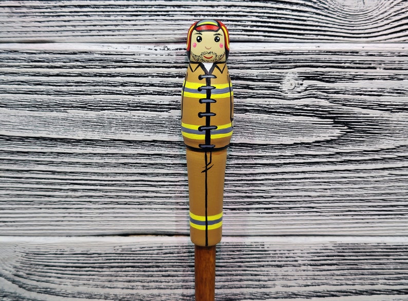 May include: A wooden figurine of a firefighter wearing a brown uniform with yellow reflective stripes and a red helmet. The figurine has a painted face with a mustache and a friendly expression.