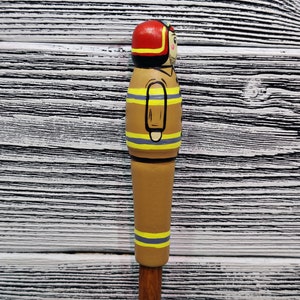 May include: A wooden figurine of a firefighter painted in brown, yellow, and red. The figurine is wearing a red helmet and has a yellow stripe down the middle of the body. The figurine is standing on a white wooden surface.