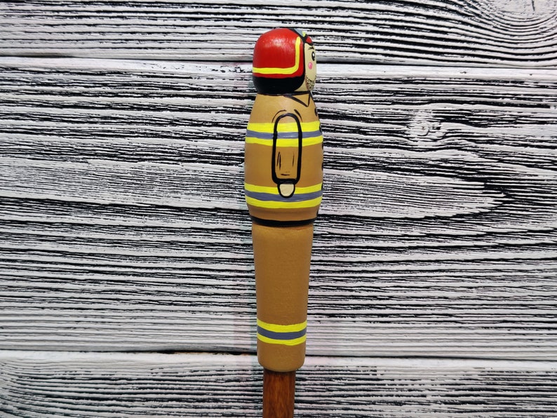 May include: A wooden figurine of a firefighter painted in brown, yellow, and red. The figurine is wearing a red helmet and has a yellow stripe down the middle of the body. The figurine is standing on a white wooden surface.