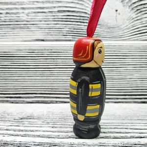 Pode incluir: Uma figura de madeira de um bombeiro vestindo um capacete vermelho com uma borda dourada, um uniforme listrado preto e amarelo e um rosto amig&aacute;vel. A figura est&aacute; pendurada em uma fita rosa.