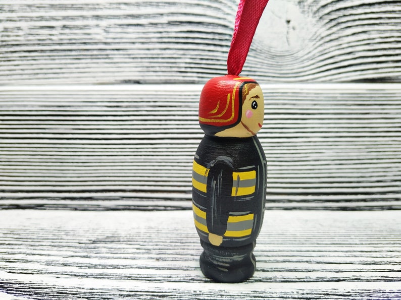 Pode incluir: Uma figura de madeira de um bombeiro vestindo um capacete vermelho com uma borda dourada, um uniforme listrado preto e amarelo e um rosto amig&aacute;vel. A figura est&aacute; pendurada em uma fita rosa.